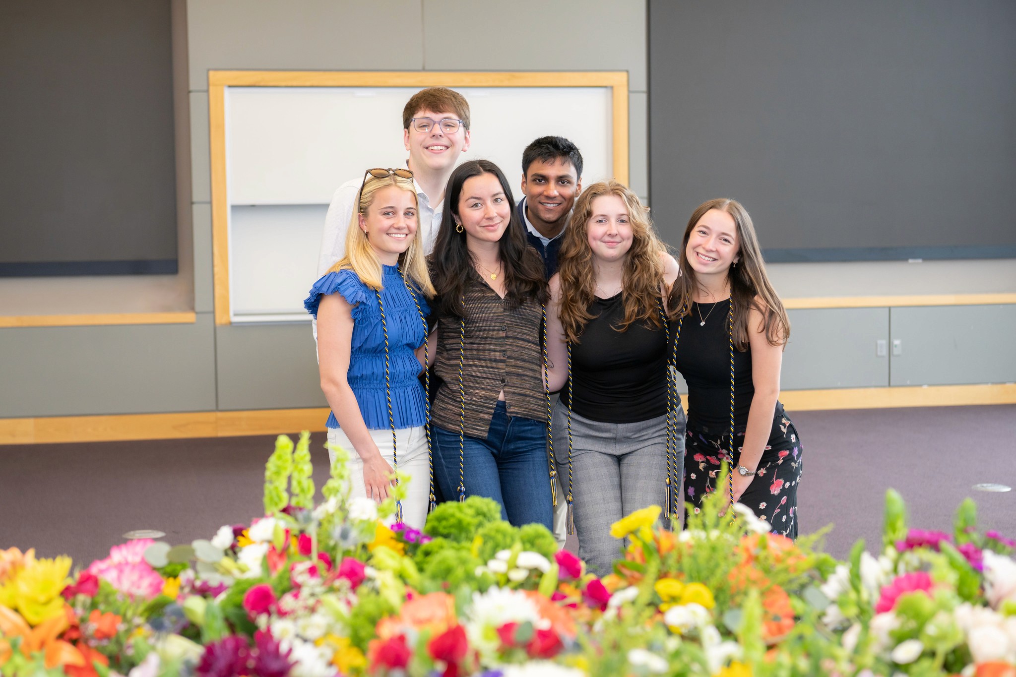 A group of six people poses together in a bright room, smiling in front of a colorful display of flowers.