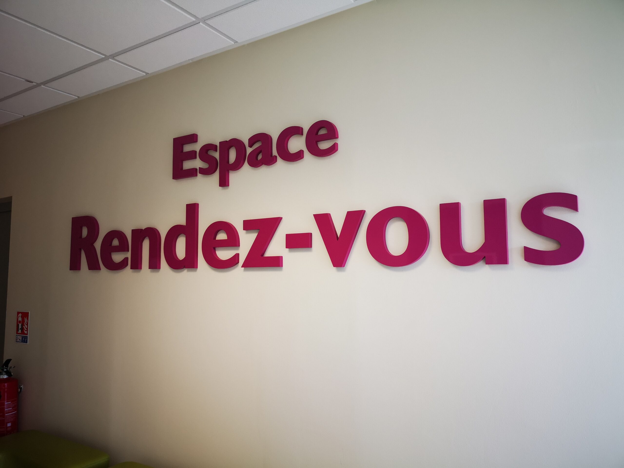 Des lettres roses vives sur un mur beige forment l'inscription « Espace Rendez-vous » dans un espace épuré et moderne. Un petit extincteur est visible à gauche.