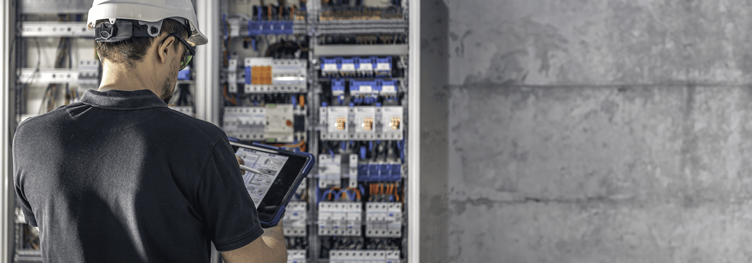 Ein Techniker mit Schutzbrille bedient das Tablet vor einem geöffneten Schaltschrank voller Kabel.