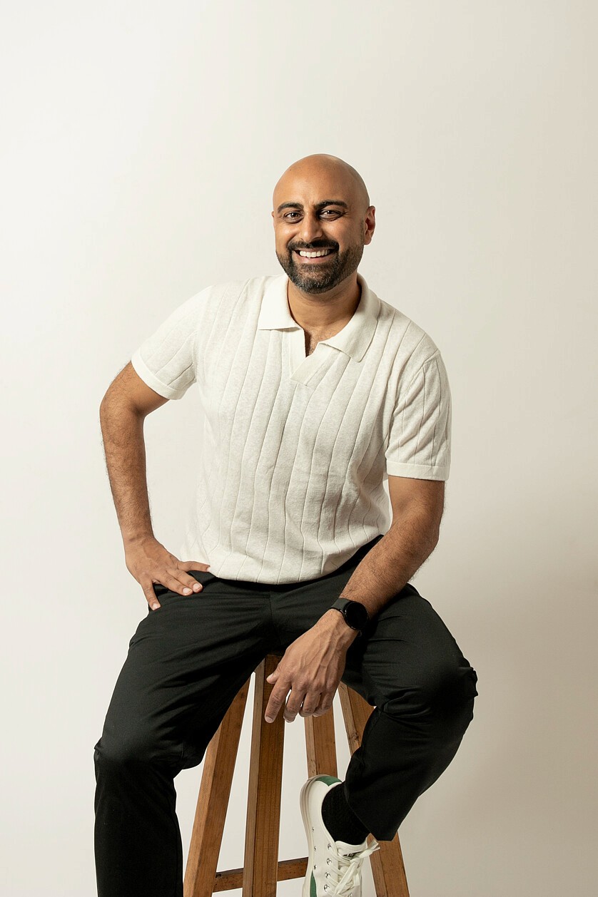 A male business strategy coach smiling confidently while sitting on a wooden stool wearing a cream shirt, black pants, and white sneakers against neutral backdrop.