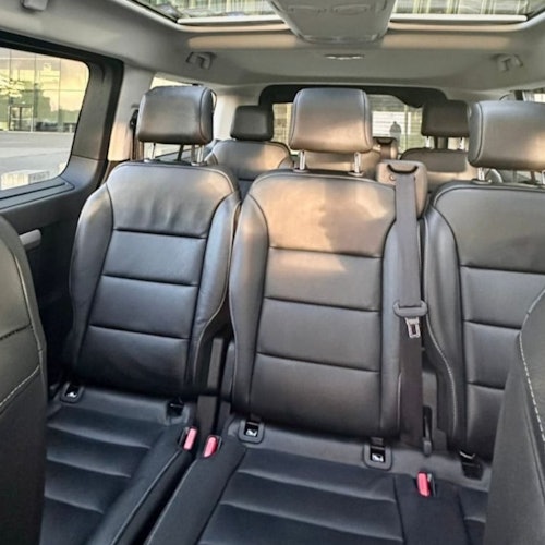 Interior de un coche con tres filas de asientos de cuero negro, cinturones de seguridad visibles y grandes ventanales.