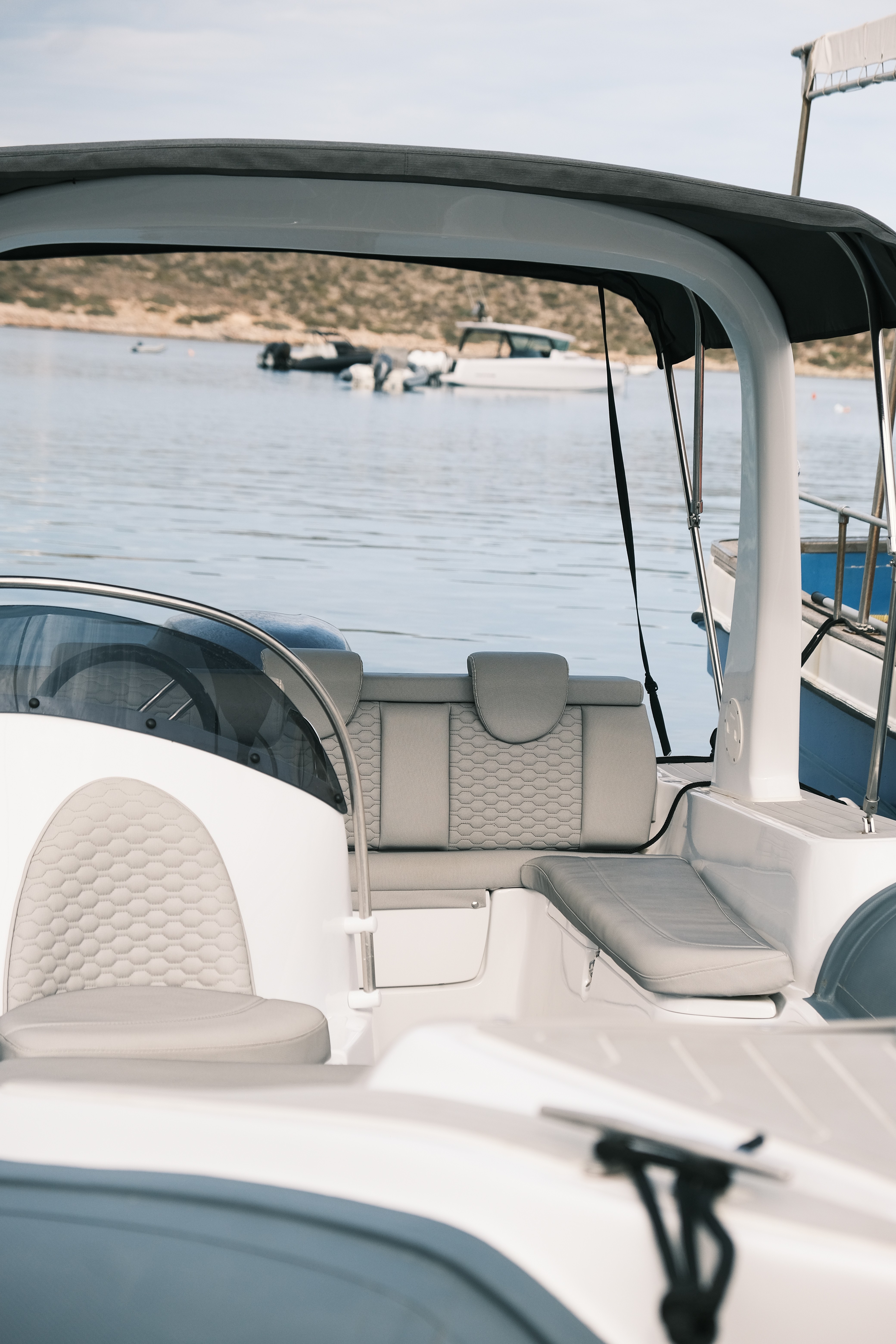 Modern yacht cockpit with white leather seating and hard top canopy, anchored in calm waters near a rocky coastline with other boats visible in the background.