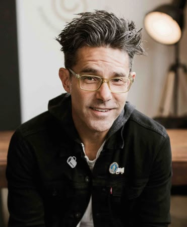 A young man with tousled hair smiles warmly while sitting in a bright, modern space.