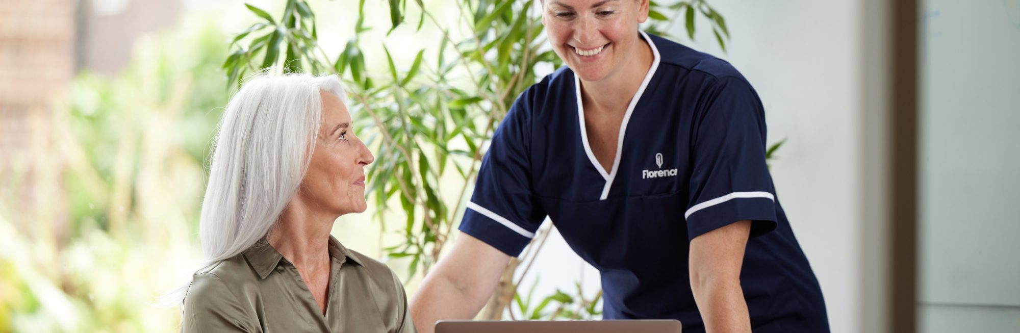 A smiling Florence nurse in navy uniform standing beside an older woman seated at a desk with a laptop, both engaged in a warm interaction