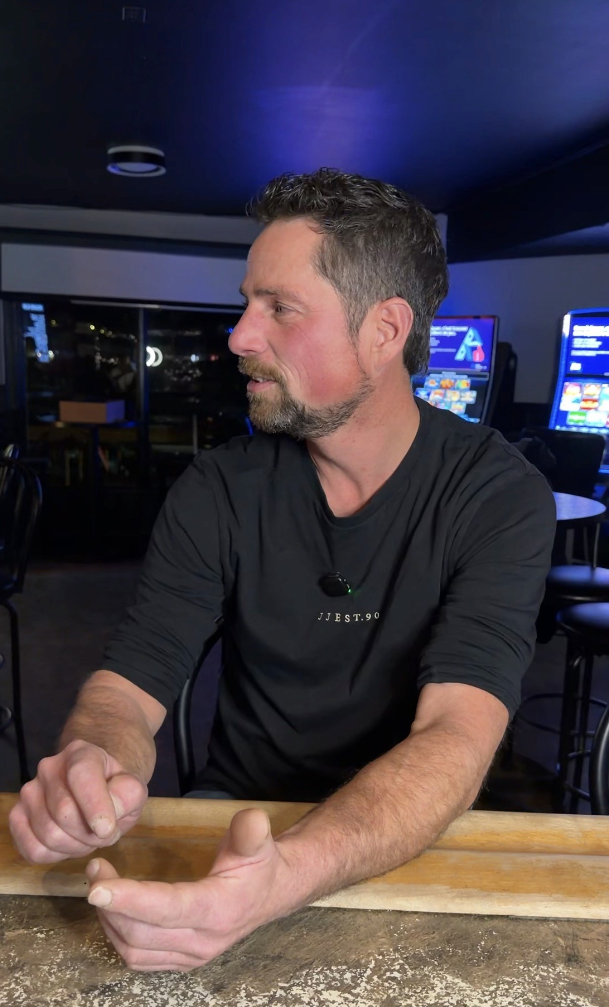 A man in a blue bomber jacket stands against a dark background. The lighting is dramatic, highlighting his focused expression. His pose is confident.