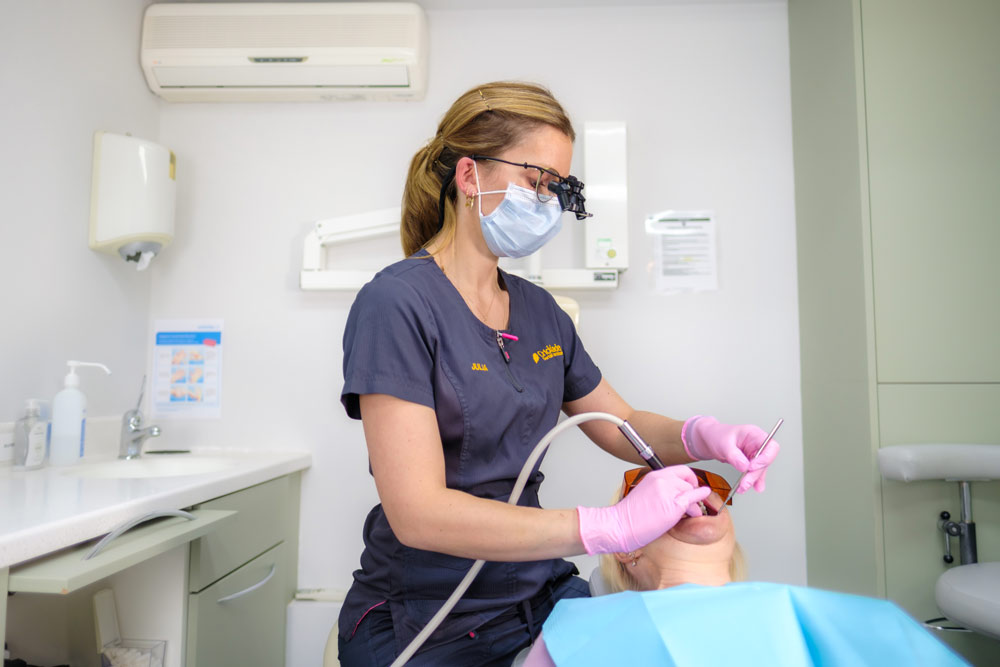 A dental professional in grey scrubs and pink gloves performs a procedure on a patient in a modern dental office, using a dental scaler while wearing a face mask and protective eyewear.