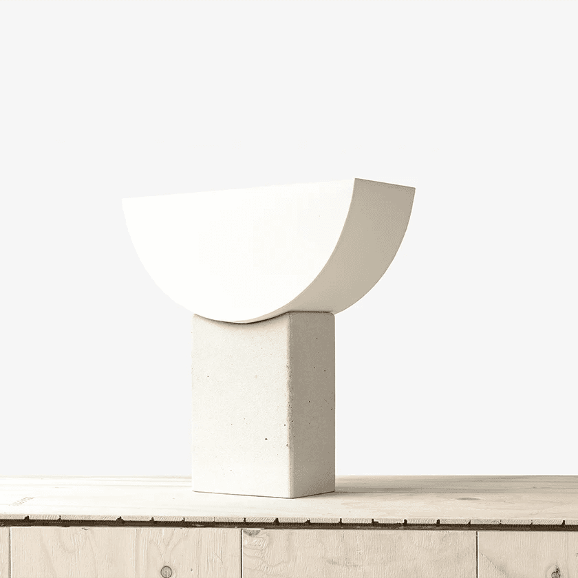 A minimalist sculpture featuring a smooth, white, semi-circular upper half resting atop a tall, rectangular concrete base, displayed on a light wooden surface.