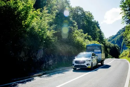 Ein weißes Fahrschulauto mit einem großen blauen Planenanhänger fährt auf einer asphaltierten Bergstraße vorbei an grünen Wäldern.