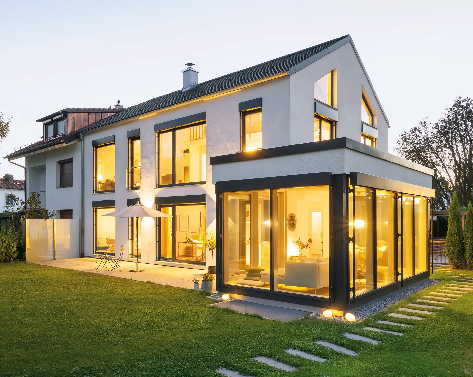 Modernes Haus mit Glasanbau und Terrasse an einem lauen Abend, beleuchtet von Innenlicht und Gartenleuchten.