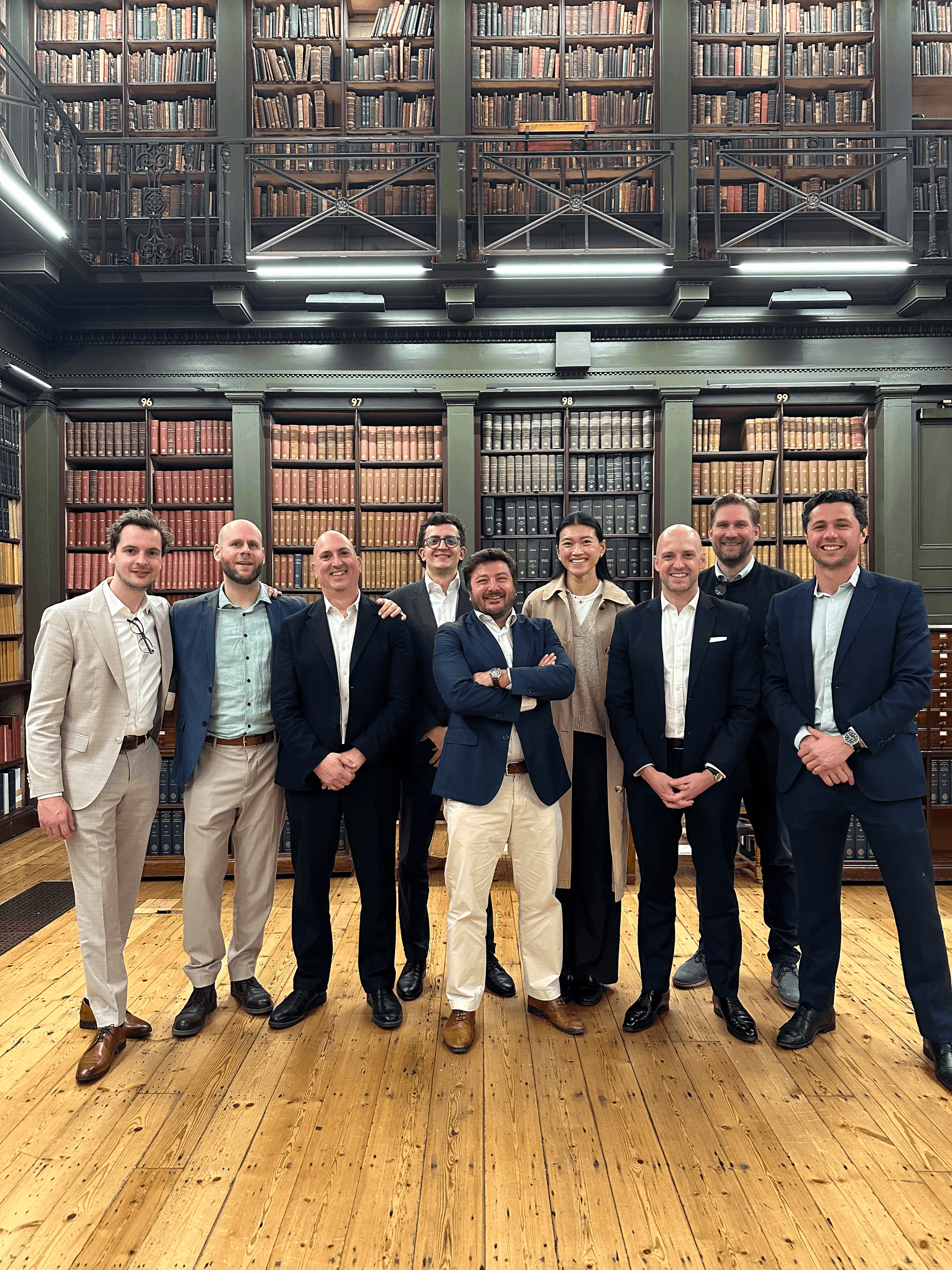 A group of seven men in formal attire standing together in a library-like setting with wooden shelves behind them.
