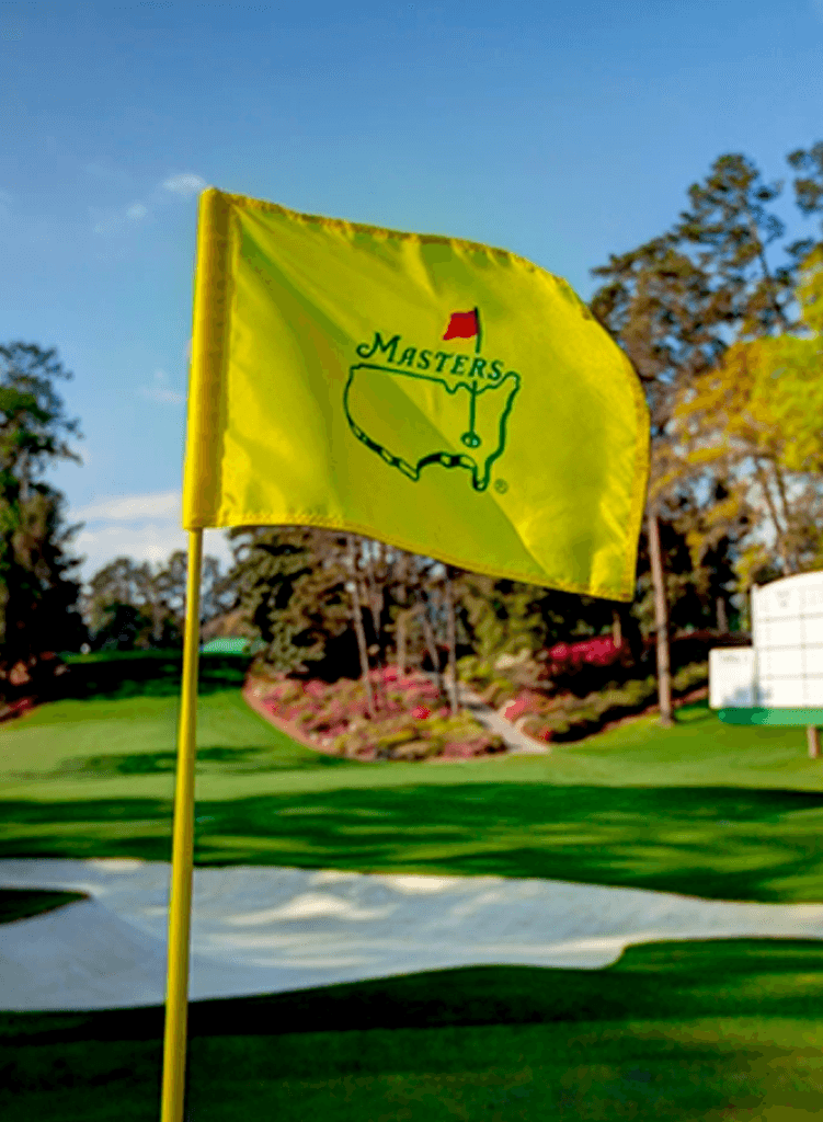 A yellow Masters golf flag flutters on a pole, marking a hole on a well-manicured green with sand bunkers, trees, and flowering shrubs in the background under a blue sky.
