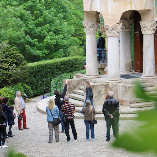 Un grupo de personas de pie frente a un edificio de piedra con altas columnas en un jardín verde y exuberante.