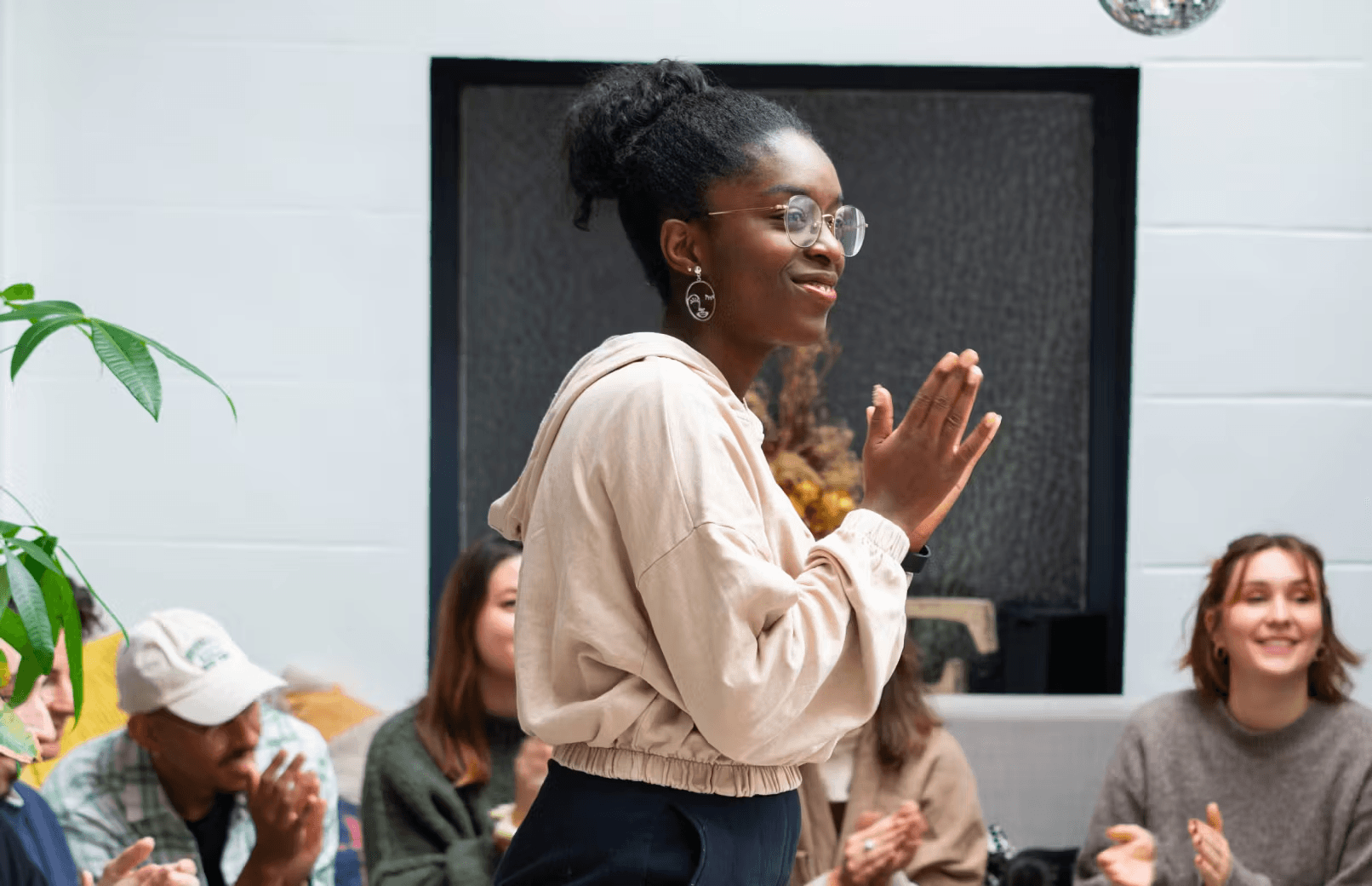 A smiling woman is standing and applauding during a friendly team meeting, surrounded by attentive colleagues in a bright space.