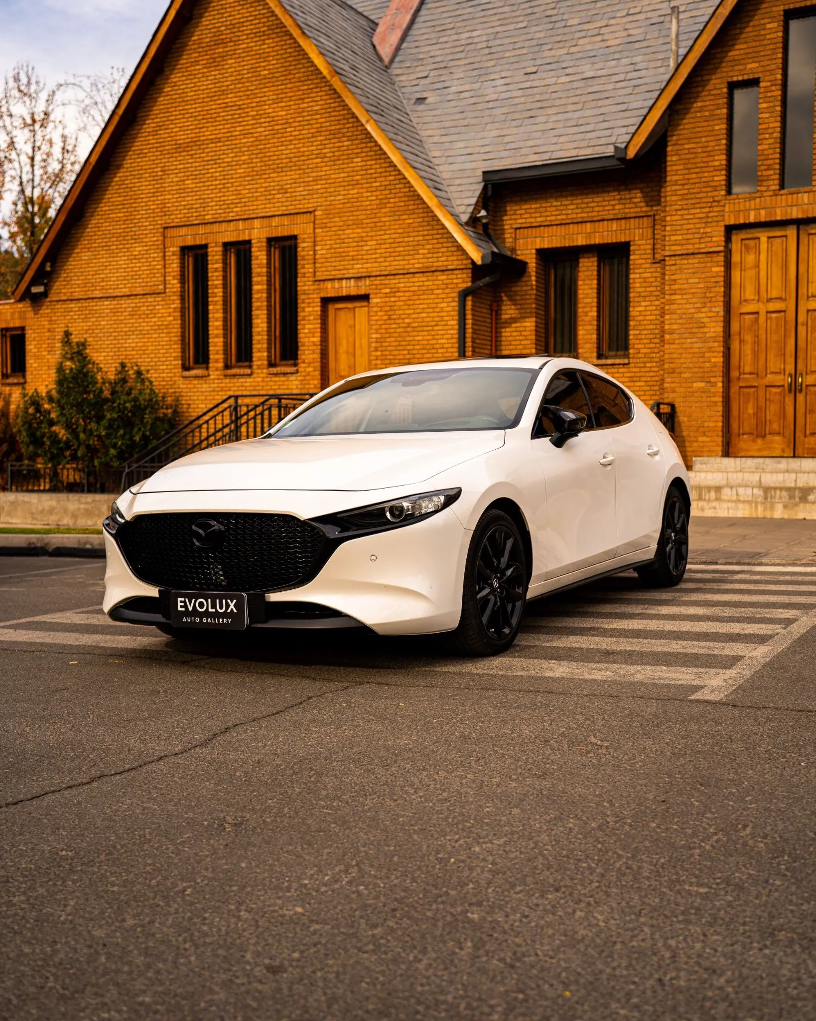 Mazda 3 blanco estacionado frente a edificio de ladrillo, vista frontal tres cuartos