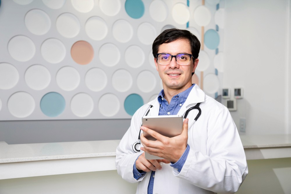 A doctor with glasses stands confidently holding a tablet, wearing a white coat with a stethoscope, in a modern office with a dotted wall pattern.