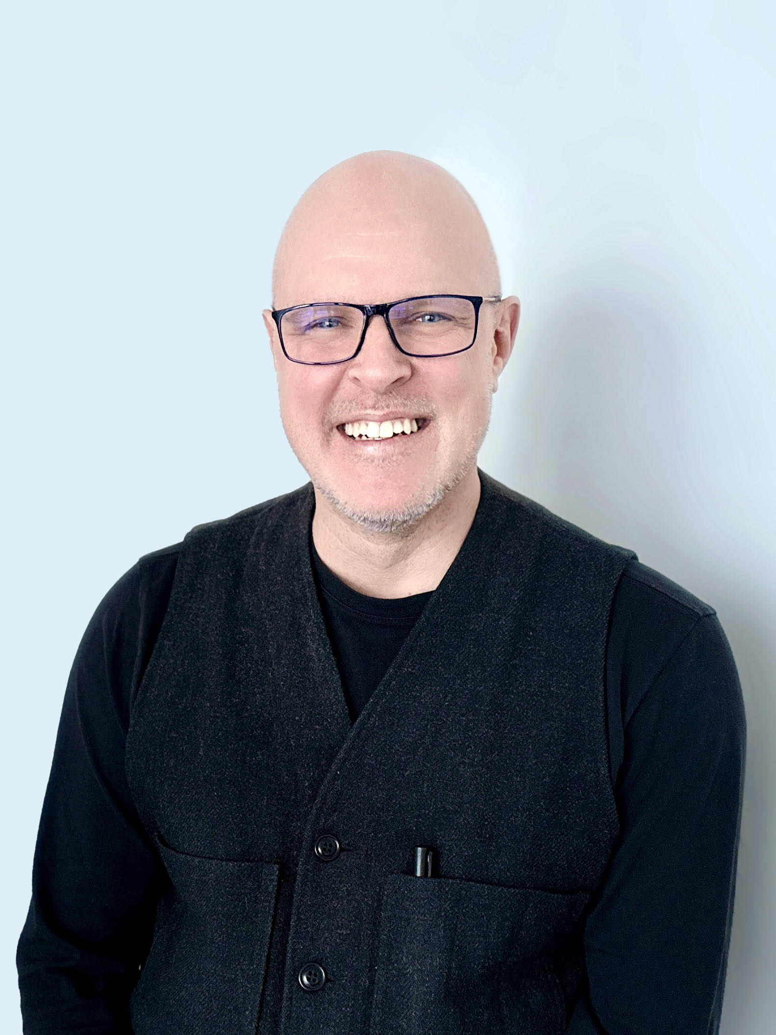 A smiling woman with her arms crossed, standing against a dark green background. She has long, dark hair.