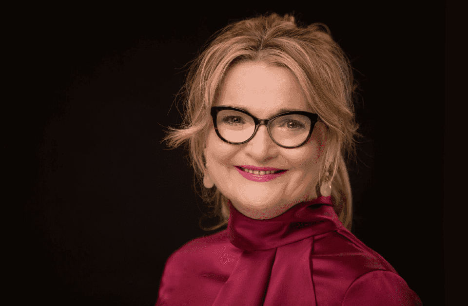Stylish woman in a pink hat and tinted sunglasses posing outdoors against a bright blue sky, wearing colorful fashion layers in warm sunlight.