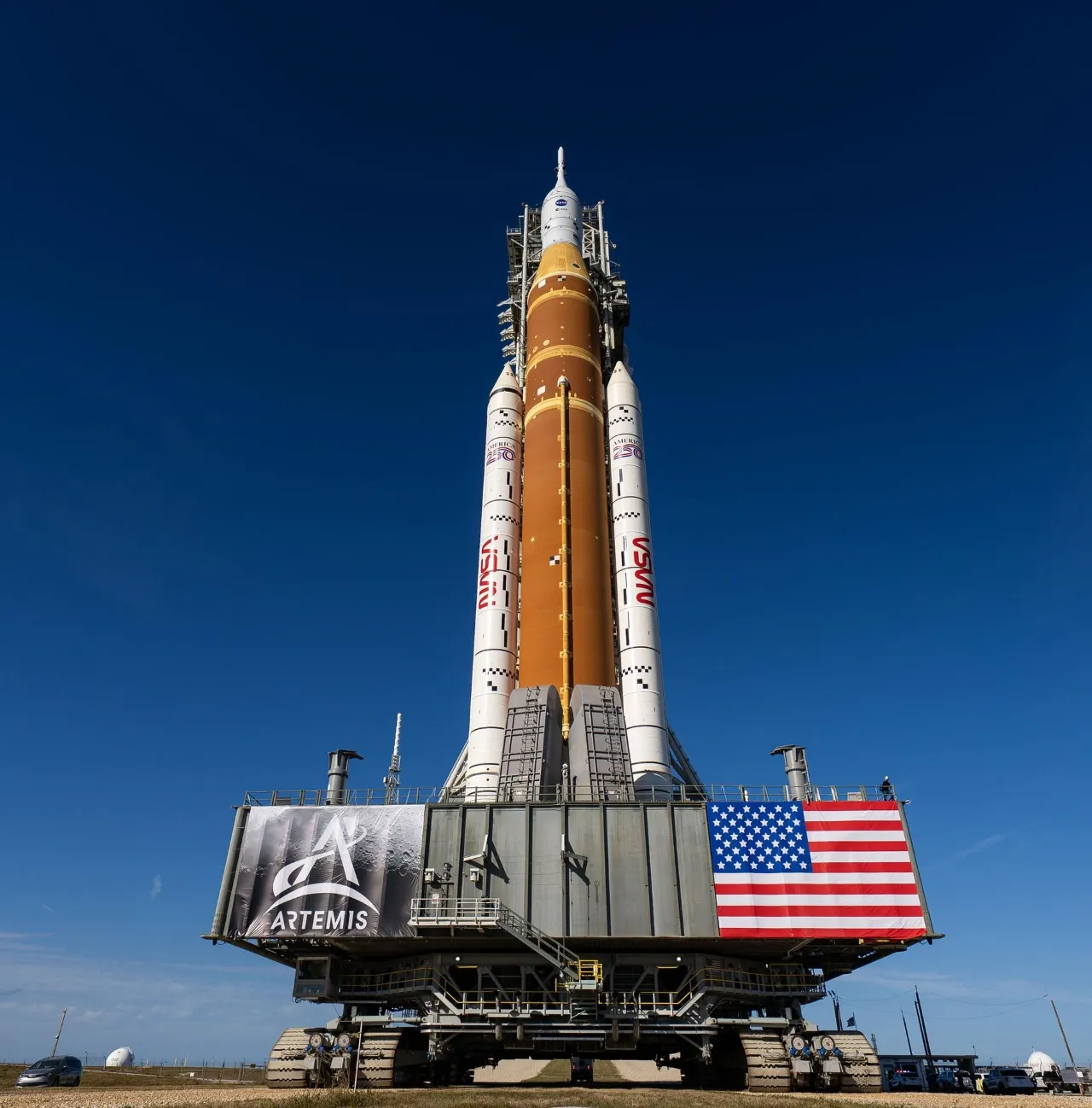 A towering space launch system rocket stands upright on a mobile launch platform under a clear blue sky, featuring a large American flag and NASA's Artemis program logo.