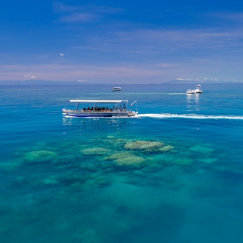 三艘船在清澈的藍水上航行,下面可見珊瑚礁,天空晴朗。最近的船上可見乘客。