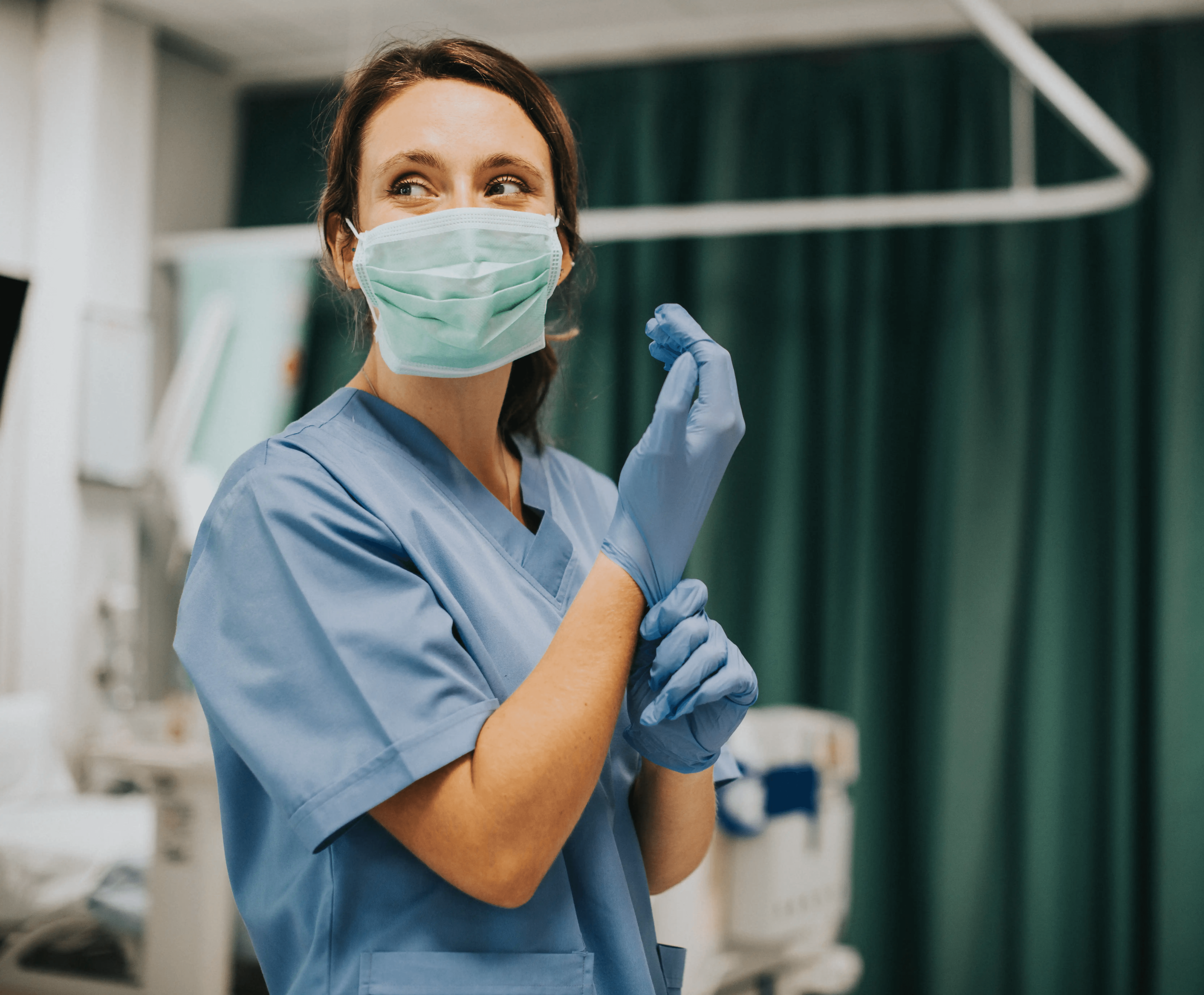 Healthcare worker in scrubs and mask putting on gloves in a medical setting.