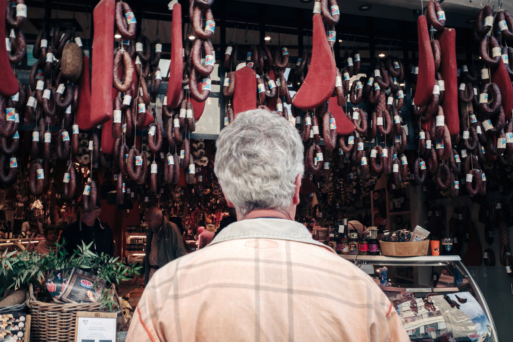 A person with gray hair stands in front of a shop displaying a wide variety of hanging cured meats and sausages, with a counter full of deli items in the foreground, creating a bustling market scene.