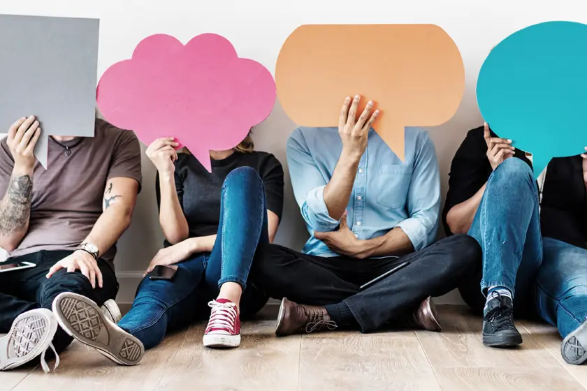 A group of people is sitting on the floor with their legs crossed. Each person holds a different colored cardboard speech bubble in front of their face, hiding their identity. The speech bubble represents a community conversation.