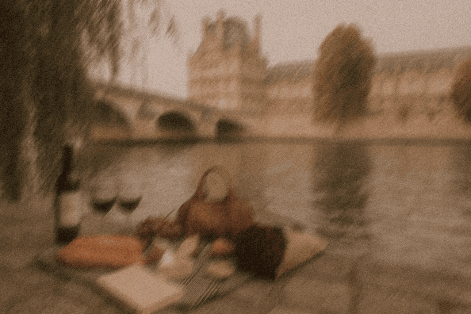 A romantic Parisian picnic scene by the Seine River, featuring wine, bread, and a handbag under the soft sepia light with a blurred view of a historic bridge and the Louvre in the background.