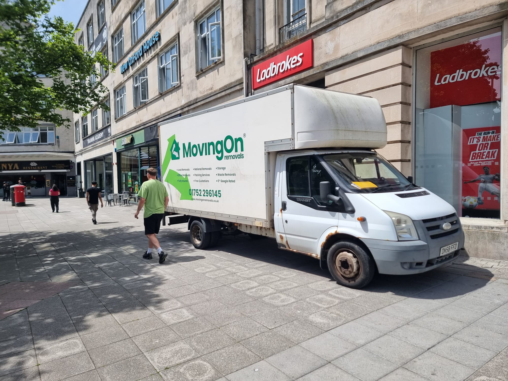 A white Moving On Removals transit van parked on a paved pedestrian street outside New Oxford House and Ladbrokes during a Plymouth city centre apartment move.