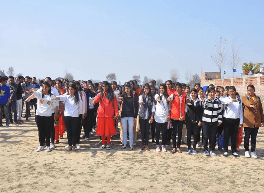 A large group of college students stands outdoors on a dusty ground, extending their arms forward to take a pledge.