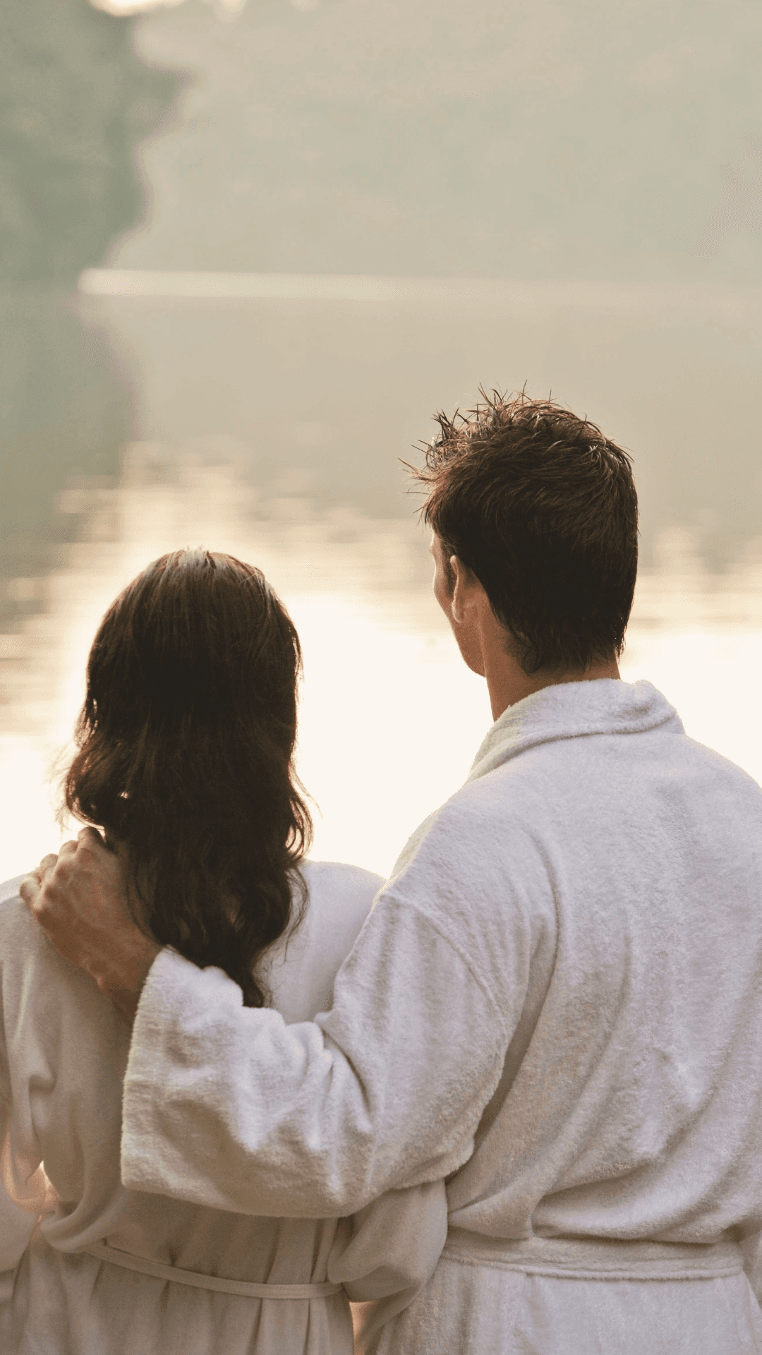 Two people in white robes looking over a tranquil lake at sunrise.