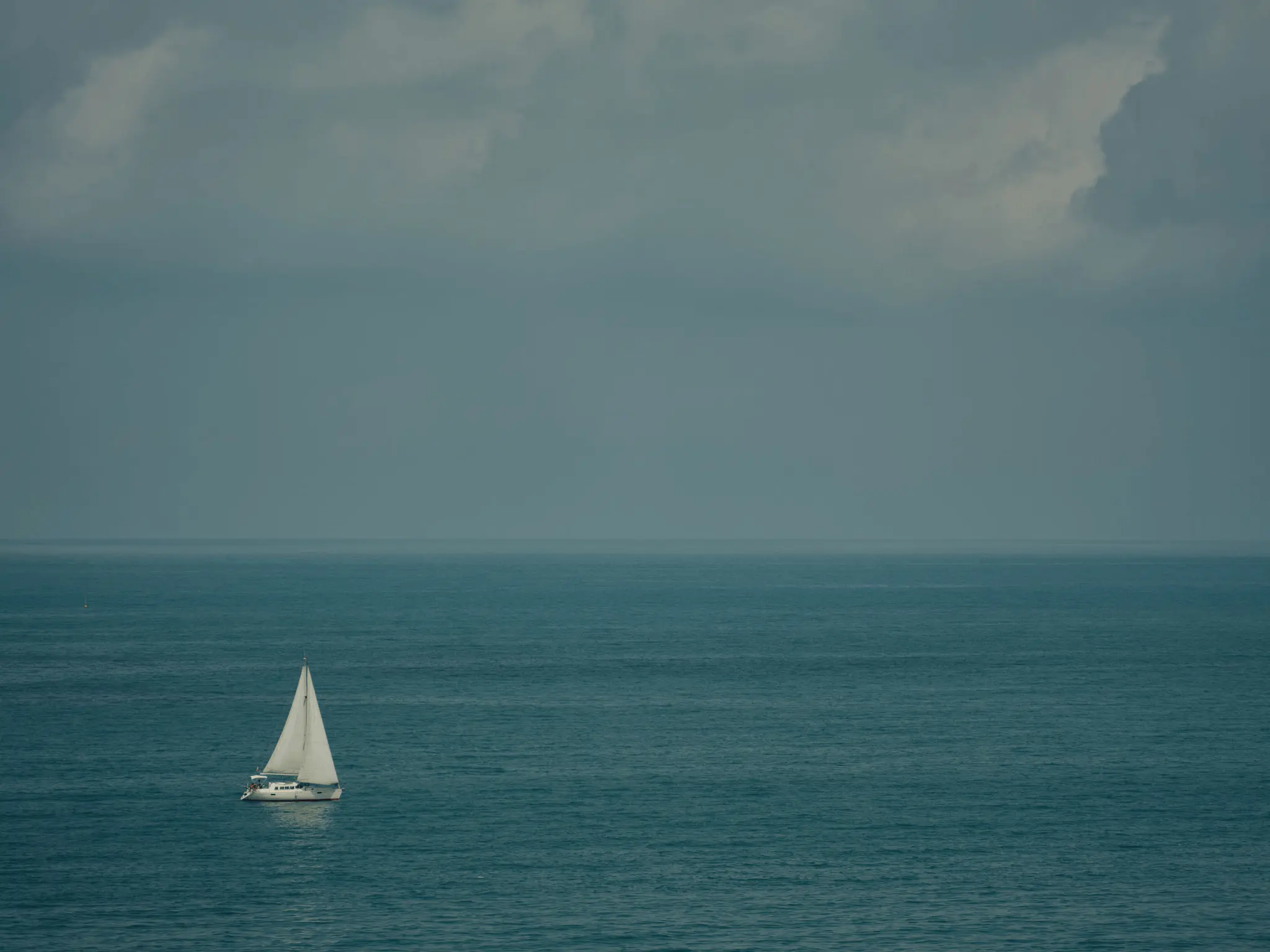 single white sailboat positioned in the lower left portion of the frame. The sailboat is peacefully gliding across a vast expanse of calm, teal-blue water. The horizon line is almost imperceptible, blending smoothly into the overcast sky above.