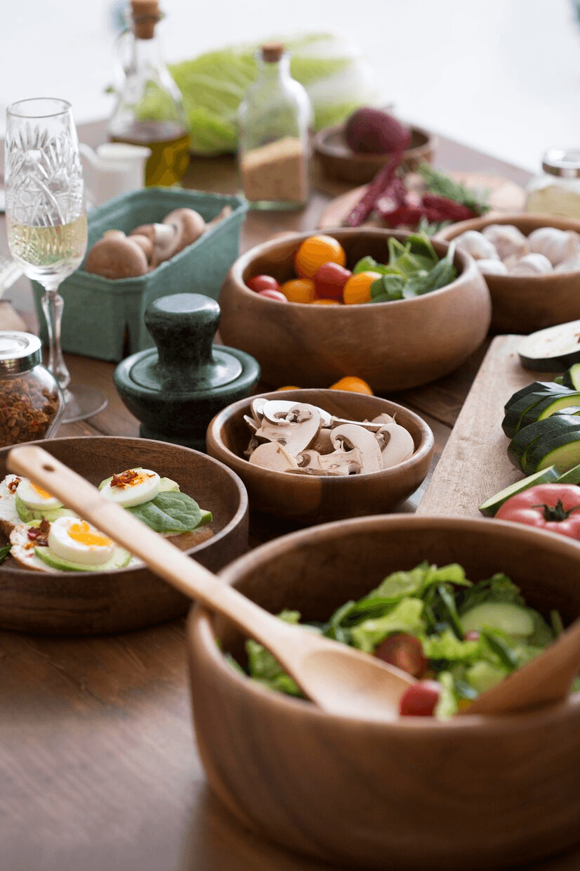 A beautifully arranged spread of Ayurvedic meals featuring fresh salads, sliced mushrooms, colorful cherry tomatoes, boiled eggs, and leafy greens, complemented by a wooden mortar and pestle on a rustic wooden table.