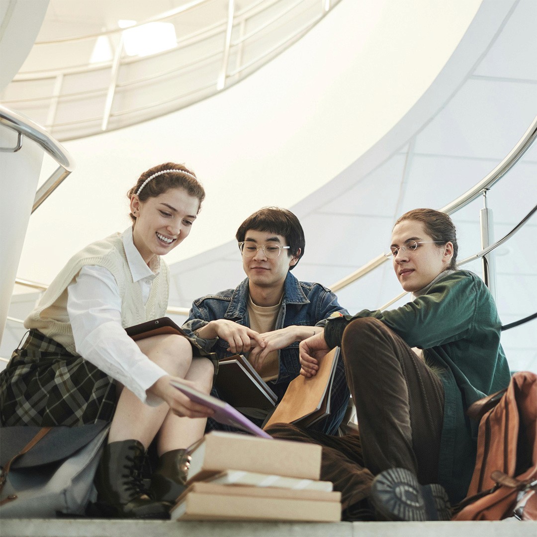 Three diverse students sitting on a university staircase all looking at the same book.