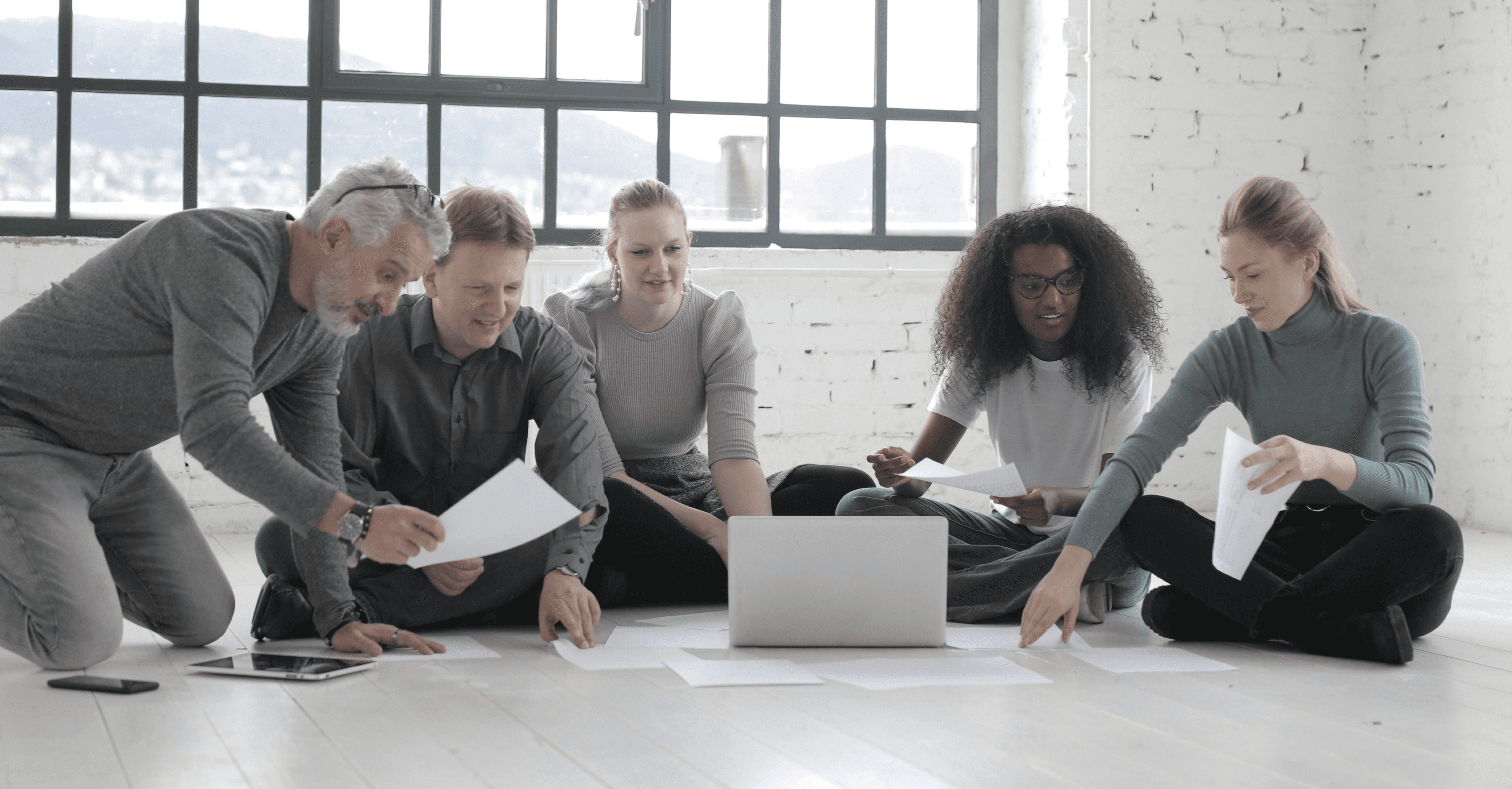 Five diverse people sit on a white floor in a bright loft, collaborating with papers and a laptop in a focused meeting.