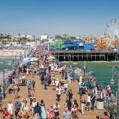 在人声鼎沸的码头上，人群熙熙攘攘。背景中有海滩、色彩斑斓的建筑物和一座摩天轮。