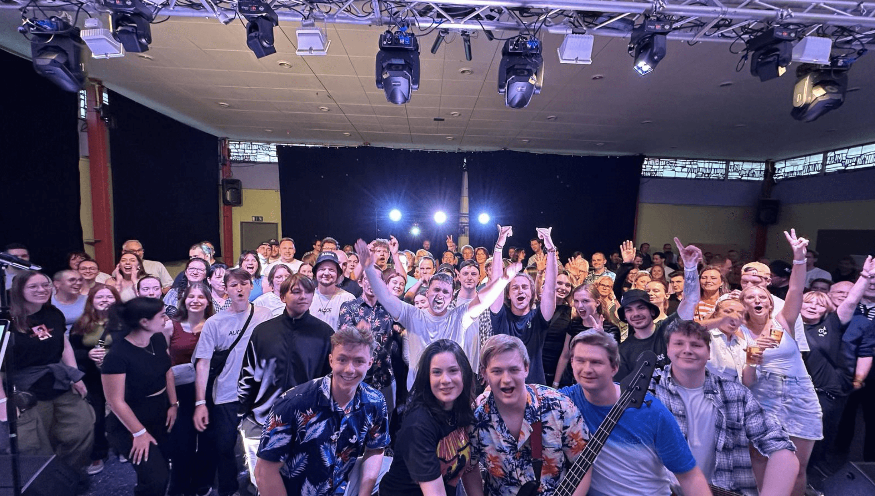 Eine Band steht auf der Bühne mit dem Rücken zum Publikum und dreht sich zur Kamera. Im Hintergrund sind Scheinwerfer und viele feiernde Menschen zu sehen. Die Stimmung wirkt energiegeladen und fröhlich.