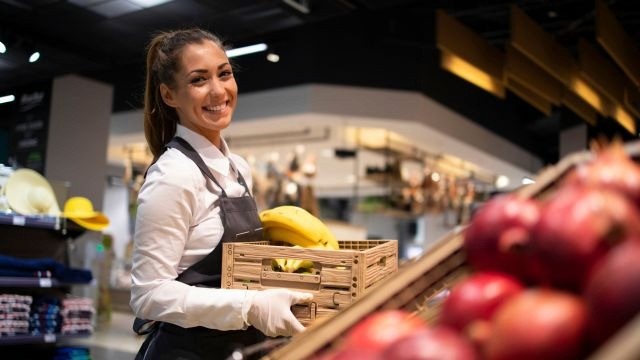 Mulher segurando caixas com frutas em corredor de mercadinho, com prateleiras e produtos ao fundo.