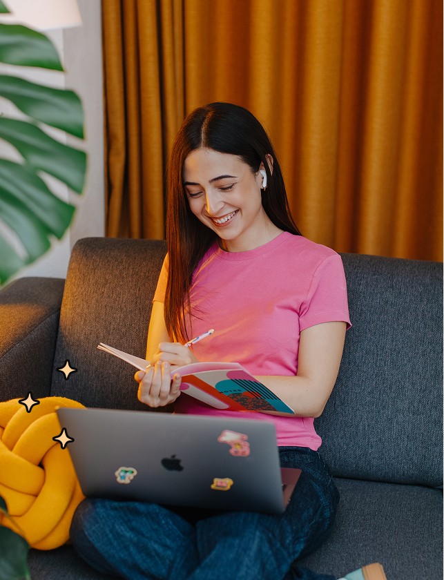 Woman smiling on a sofa, writing in a notebook with a stickered MacBook on her lap.