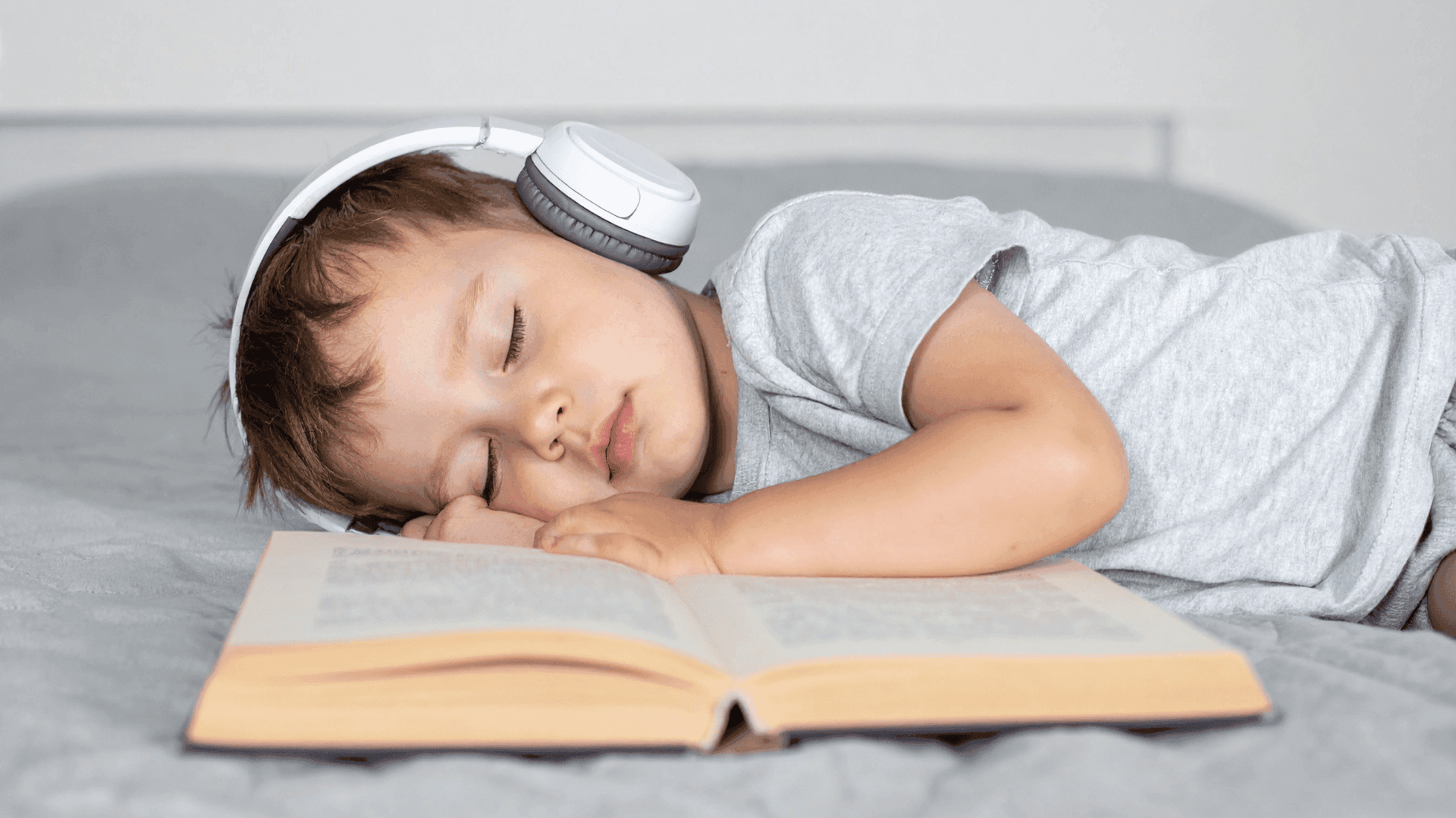 A boy asleep on a bed, wearing headphones with a book in front of him.