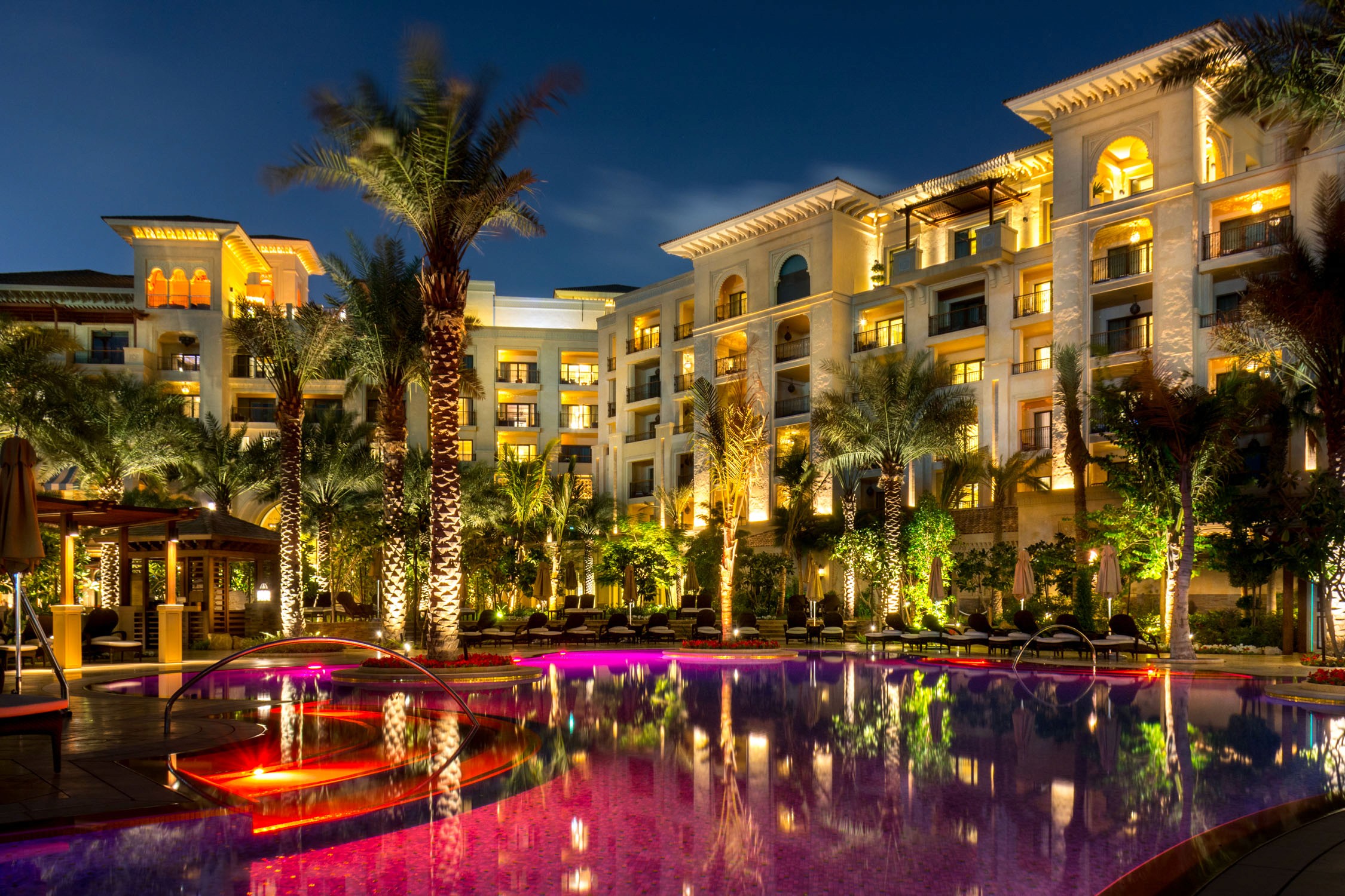 A luxury resort hotel with lit balconies and palm trees overlooks a swimming pool illuminated with pink and purple lights at
