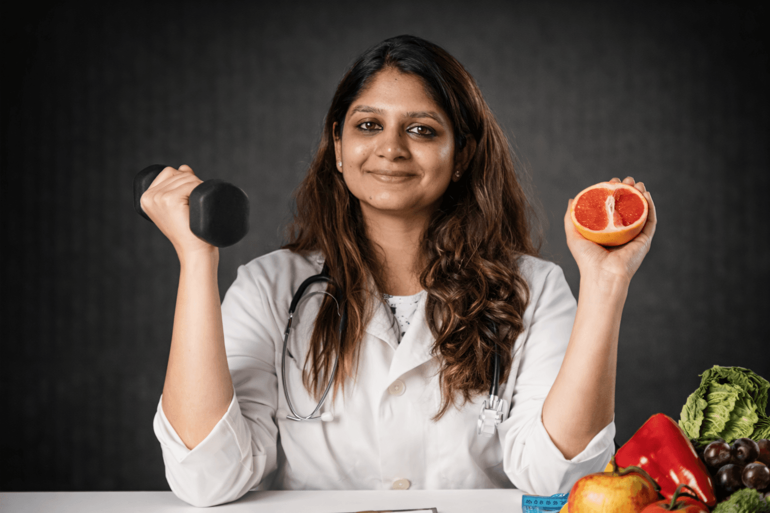 A woman in a white coat, with a stethoscope around her neck, holds a dumbbell in one hand and half a grapefruit in the other. On the table are fruits, vegetables, and a blue measuring tape, symbolizing her wellness-focused nutrition plan. She smiles at the camera.