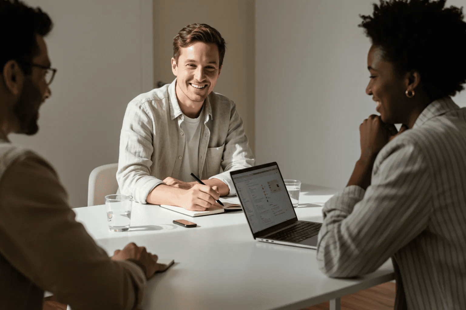 Three colleagues sitting around a table, smiling and discussing work with laptops open in front of them during a team meeting.