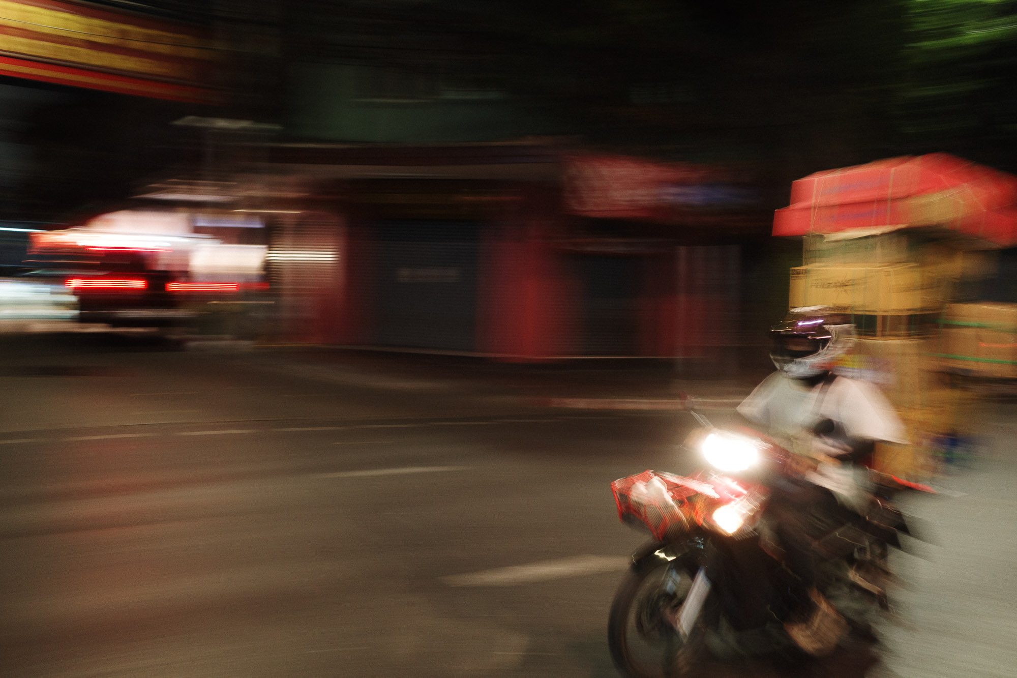 A person wearing a helmet rides a motorcycle through a dimly lit urban street at night, with motion blur capturing the sense of speed and a background of a softly illuminated building facade.