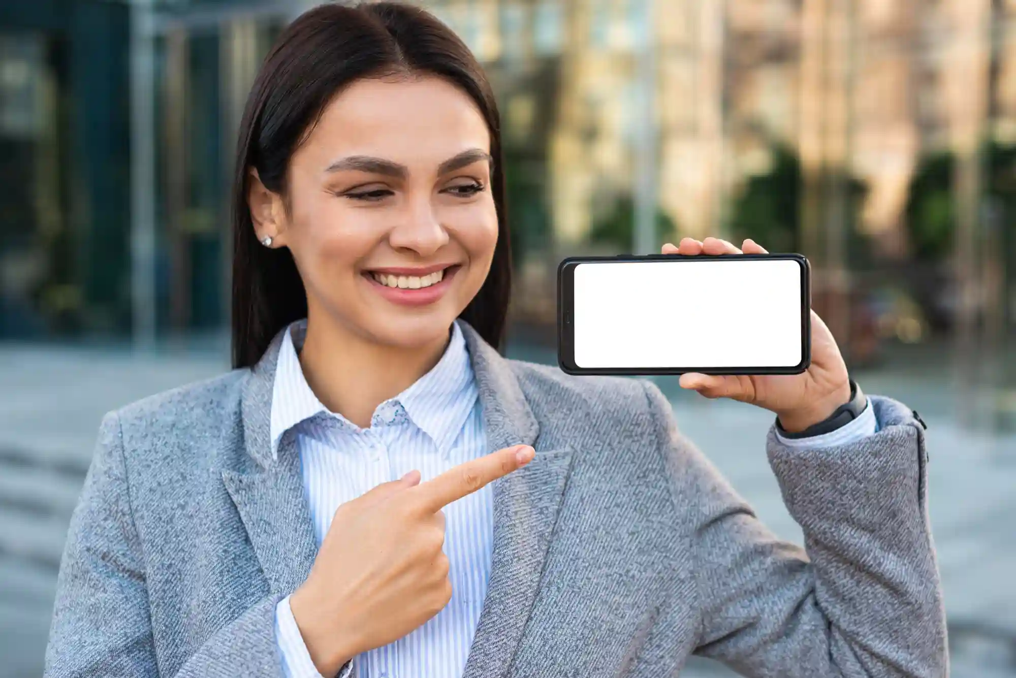 Smiling businesswoman pointing to a blank horizontal smartphone screen, perfect for showcasing your latest mobile app or digital service.