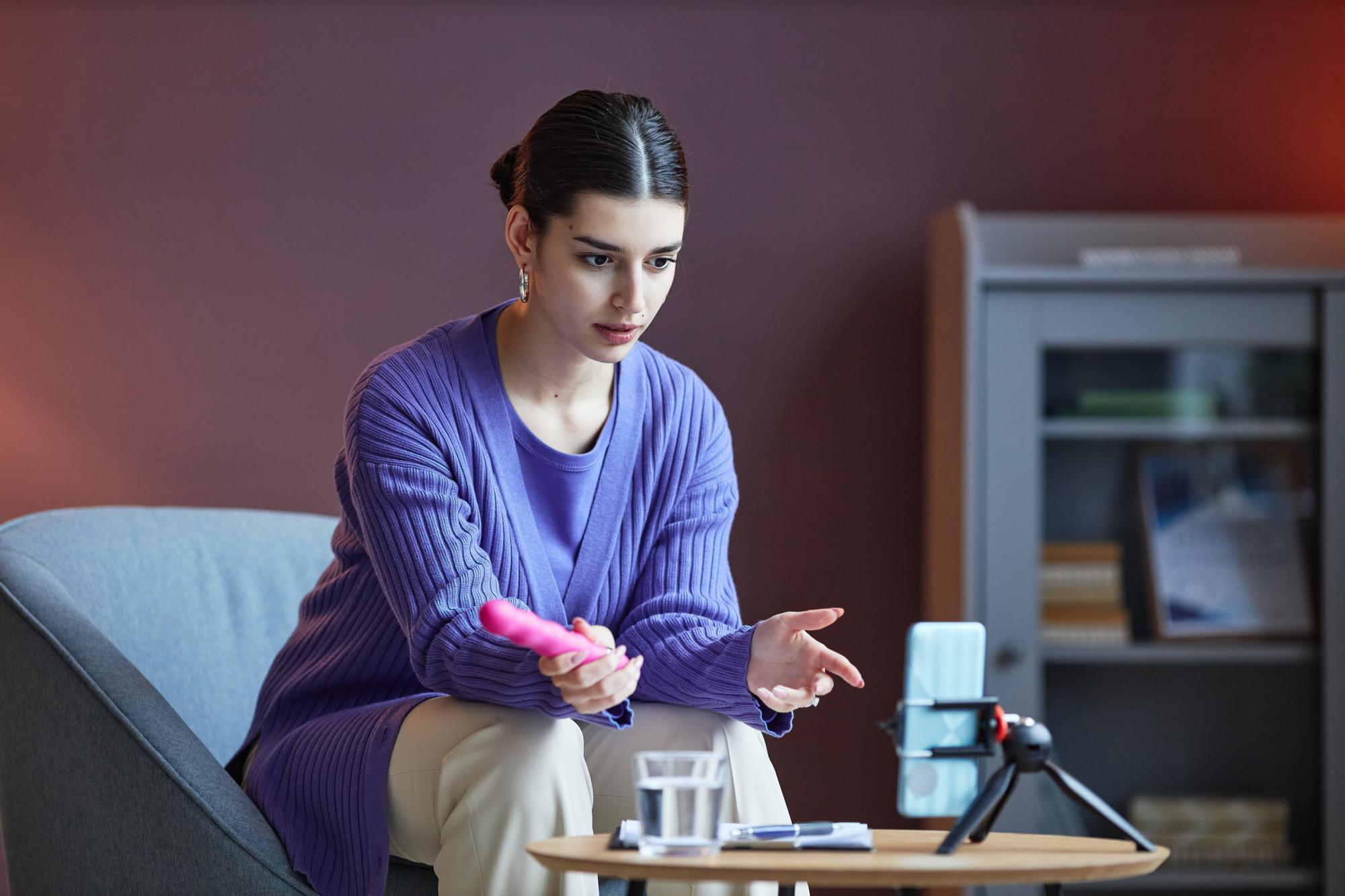 A woman seated on a couch, holding a phone in one hand and a remote control in the other, appears relaxed and engaged.