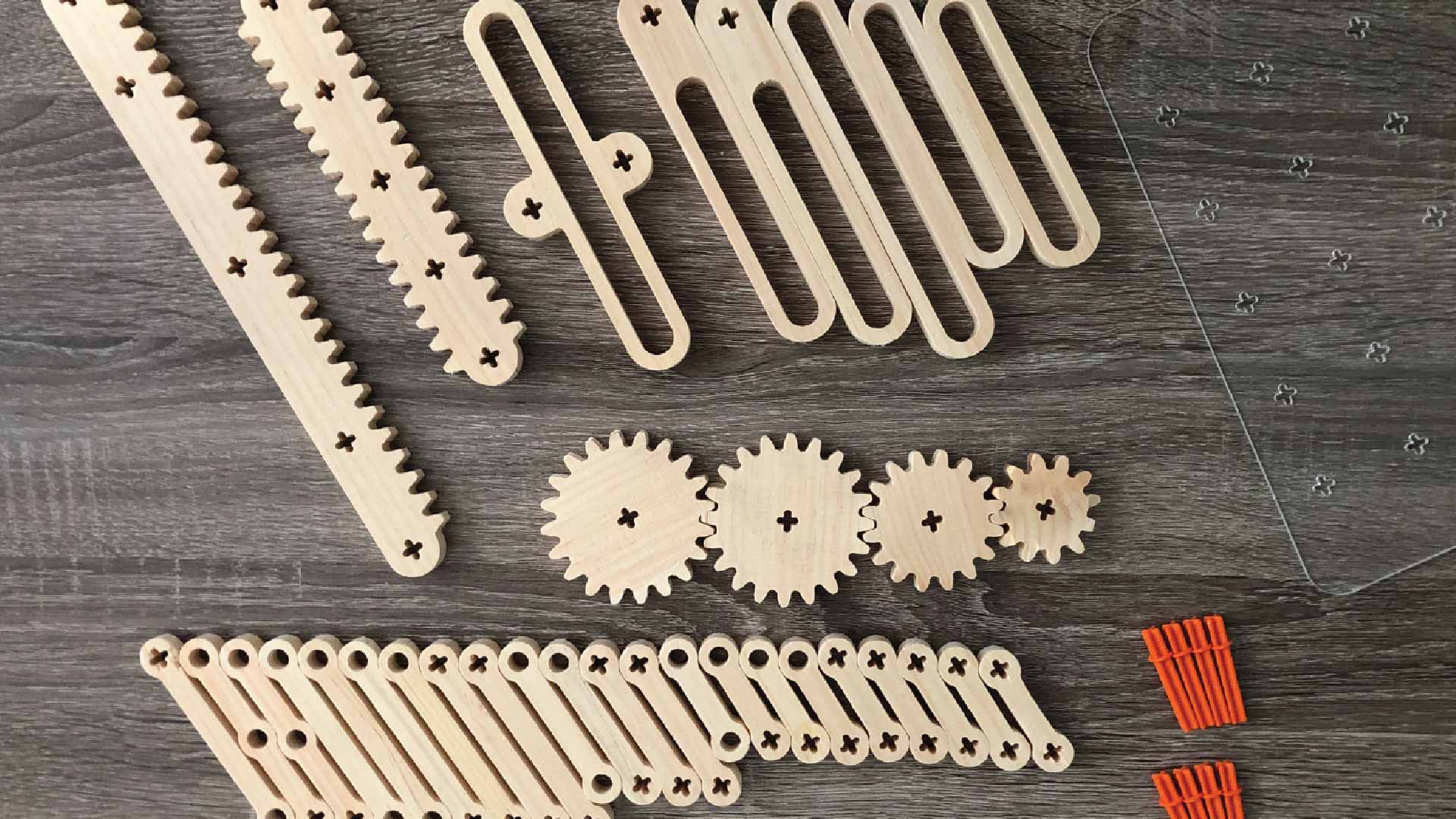A wooden gear set is arranged on a dark gray wooden surface. The set includes several gear shapes with small holes and dark markings, arranged in groups. Near the bottom right corner, there are two stacks of orange plastic clamps. A clear plastic ruler with small holes is positioned diagonally across the upper right portion of the image.