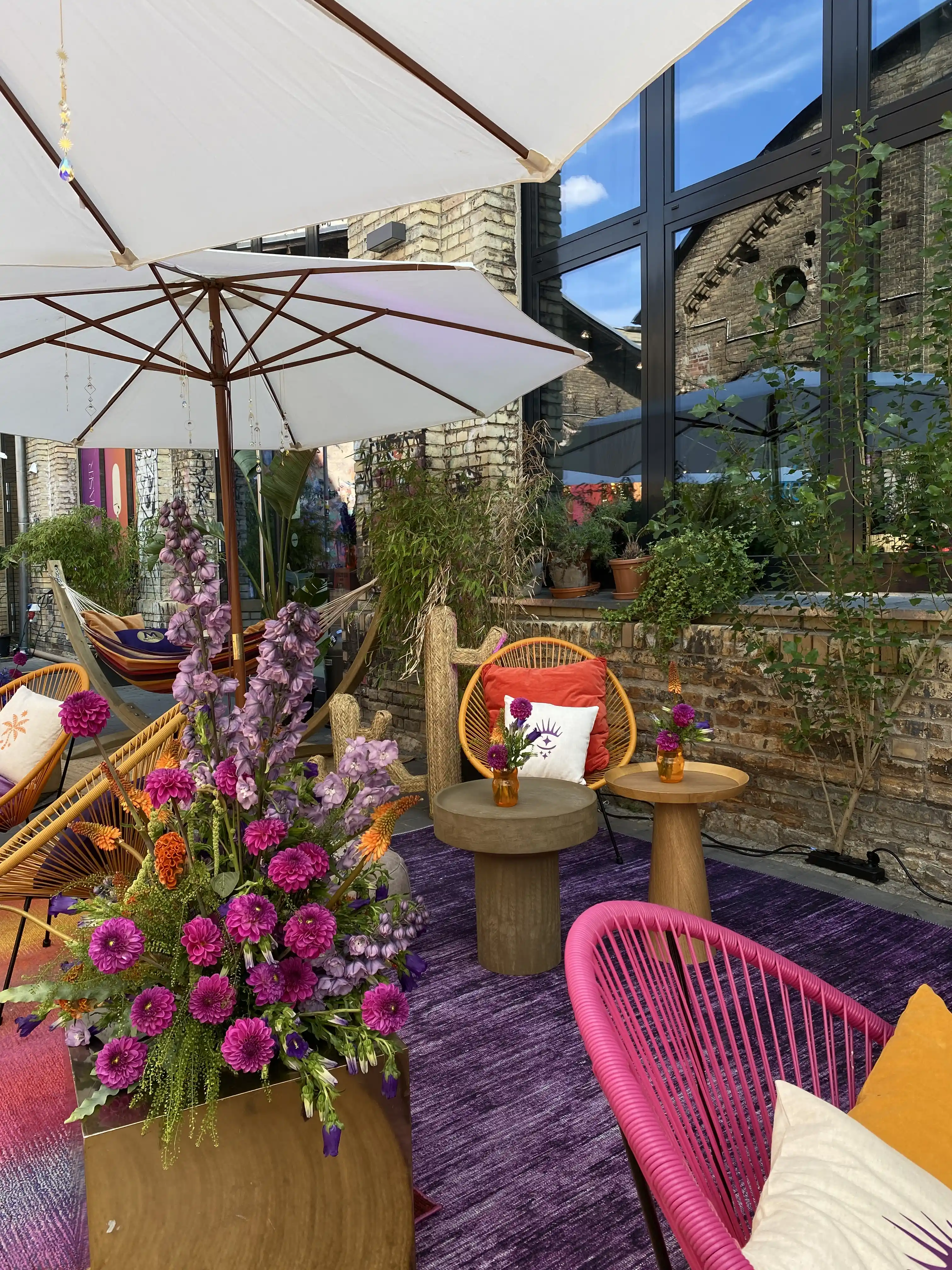 Lounge corner with lush flower arrangement, pink wire chairs, wooden side tables and parasol.