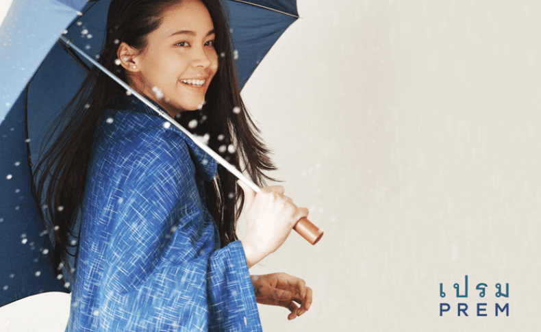 A person with short dark hair and light skin is wearing a dark navy blue poncho with a zipper closure, standing against a plain off-white background. Their gaze is directed to their left with a neutral expression.