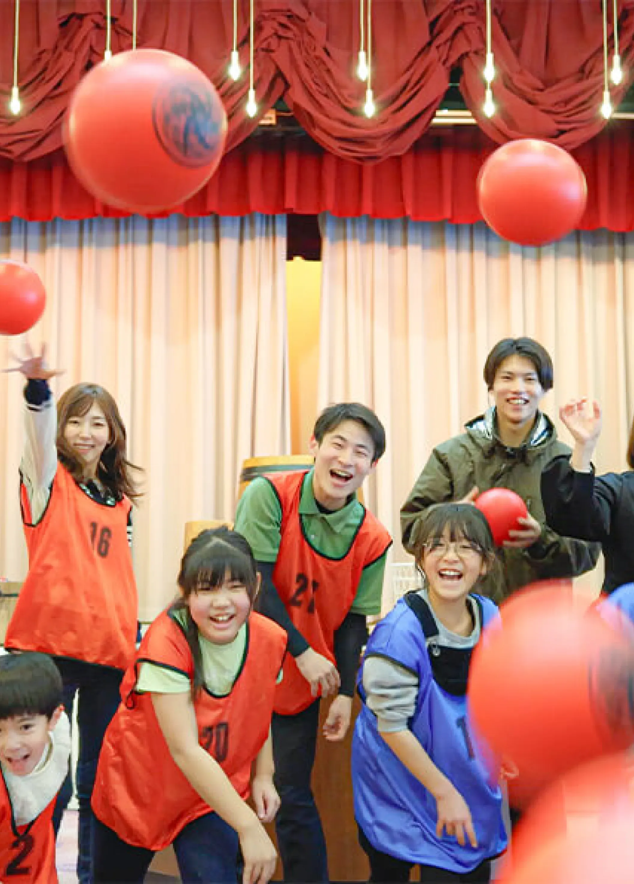 A joyful and energetic group of children and adults from our kids academy laughing together during a lively ball-tossing game. Participants are wearing red and blue pinnies in front of a stage with a red curtain, with several red balls caught in mid-air.