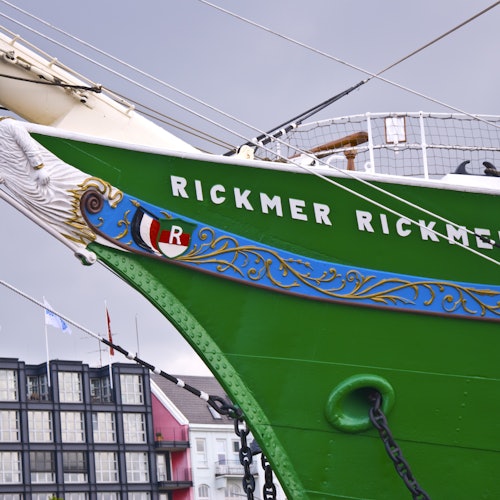 Bow of a green ship named "Rickmer Rickmers" with decorative blue and gold trim, docked near buildings under a gray sky.